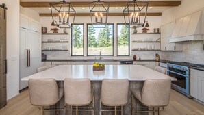 A modern kitchen with a large island, bar stools, pendant lighting, and a view of trees through large windows. - Breathtaking Big Sky Luxury Retreat (Big Sky)