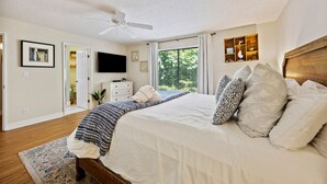 Une chambre à coucher comprenant un lit, un ventilateur de plafond, une télévision, une table de chevet et une fenêtre avec des stores.