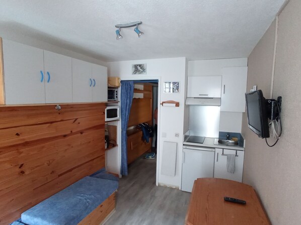 A compact room with a wooden bench, a kitchenette, and a small TV. - Chaleureux Studio , Parking en Centre Ville (Cauterets)