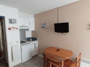 A compact kitchen with white cabinets, a small sink, and a dining area with a wooden table and chairs. A television is mounted on the wall. - Chaleureux Studio , Parking en Centre Ville (Cauterets)