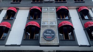 A building with multiple red awnings labeled "BABILON" and a central sign reading "THE BABILON Suites".