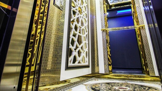 Interior de un ascensor con puerta de diseño geométrico, paredes metálicas y suelo reflectante.
