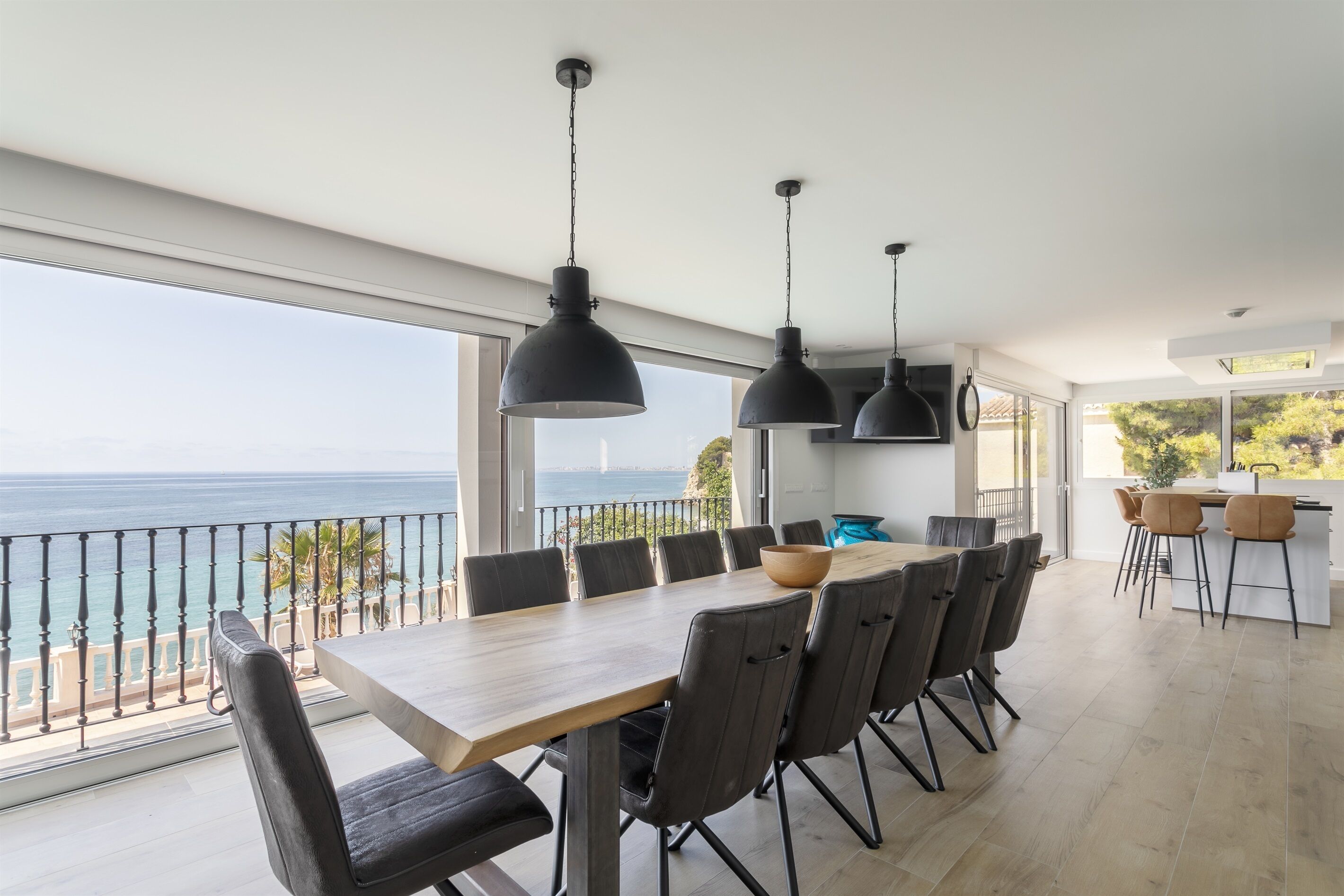 Een moderne eetruimte met een grote tafel en stoelen, een uitzicht op zee en hanglampen.