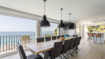 Een moderne eetruimte met een grote tafel en stoelen, een uitzicht op zee en hanglampen.
