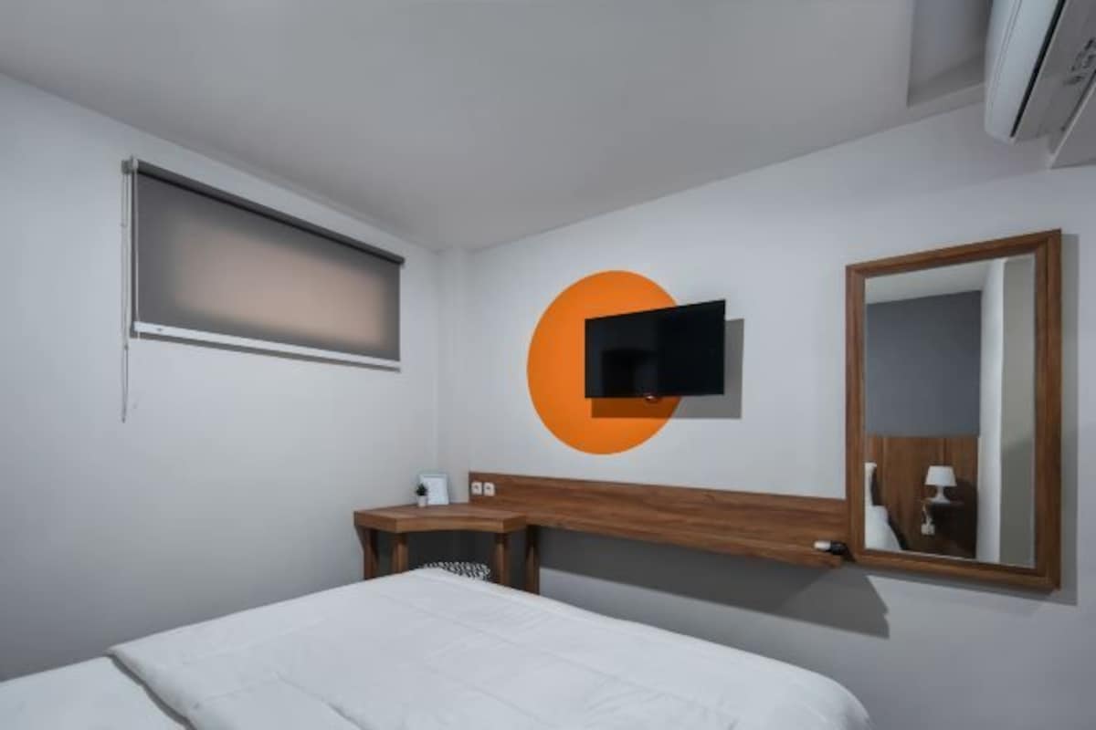 A hotel room with a bed, a desk, a TV mounted on the wall, and a window with blinds.
