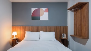 Ein Hotelzimmer mit einem hölzernen Kopfteil, einem Bett mit weißer Bettwäsche, einem Nachttisch mit Lampe, einem kleinen Tisch mit einem Kaktus und einem Holzregal.