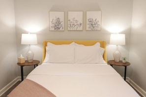 A bed with a yellow headboard, two bedside tables with lamps, and three framed floral artworks on the wall.