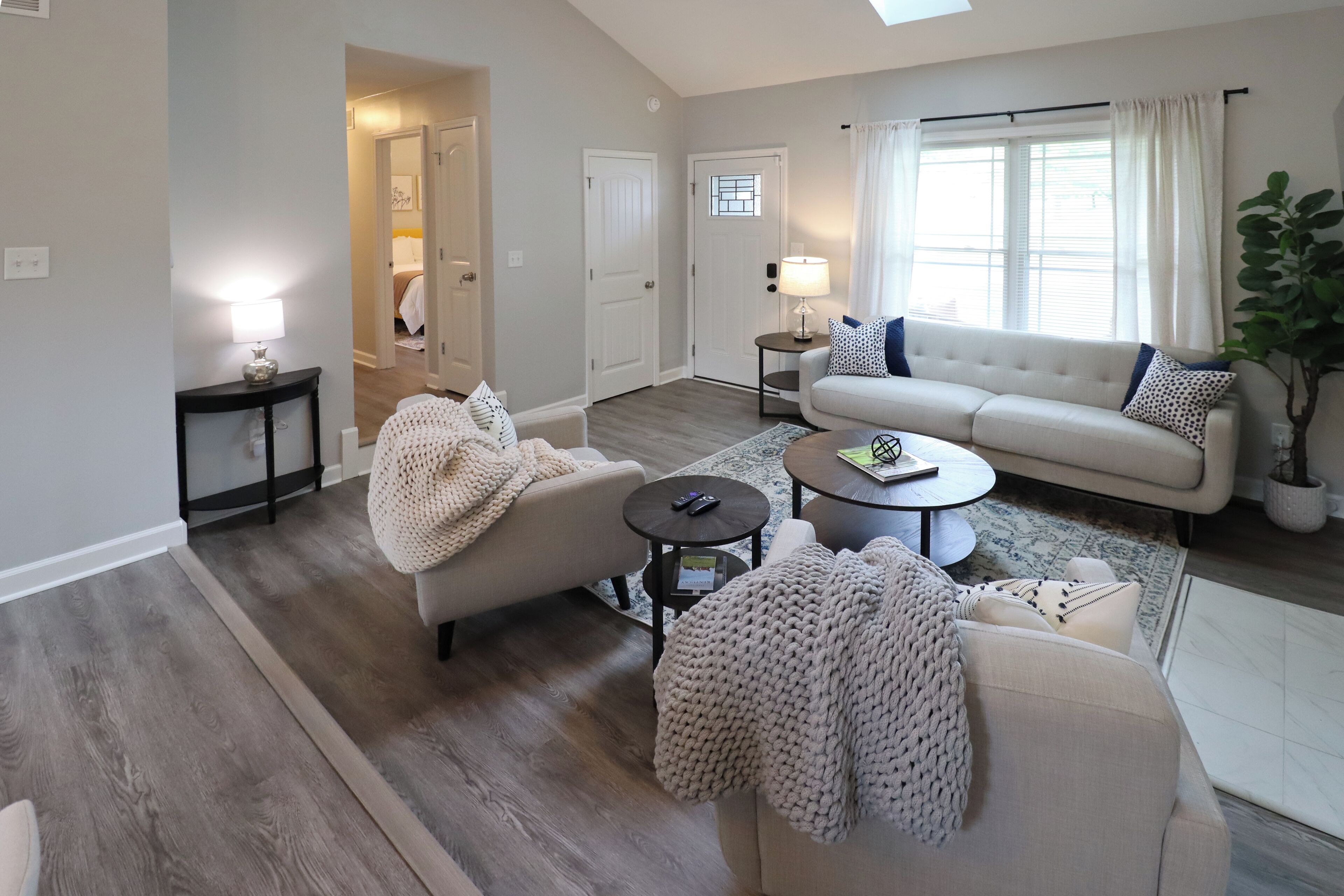 A living room with a sofa, armchairs, a coffee table, and a rug. There are two doors, one leading to a bedroom and another to a hallway.