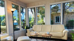 A sunlit room with a beige sofa, a round wooden coffee table, and a matching ottoman. Large windows offer views of a garden and a brick building.