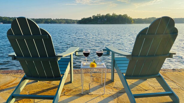 Two Adirondack chairs facing a lake with a table between them, holding a glass of red wine and a yellow flower.