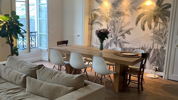 A living room with a beige sofa, a dining table with chairs, a large window, and a tropical mural on the wall. - Très bel Appartement Familial Parisien aux Pieds des Invalides (Paris)