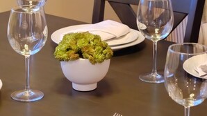 A dining table set with white plates, silverware, and clear wine glasses. A white bowl with green foliage is placed in the center. - Tranquil Coastal Retreat! (Parksville)