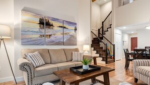 A spacious living room with a beige sofa, a wooden coffee table, and a large painting of a sunset over a lake. - Tranquil Coastal Retreat! (Parksville)