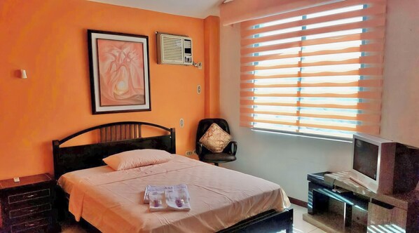A bedroom with an orange wall, a bed with a pink bedspread, a chair, a TV on a stand, and a window with blinds.