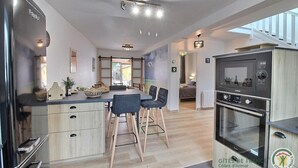 A modern kitchen with a refrigerator, oven, and microwave. There are two bar stools and a dining table. The room has wooden flooring and white walls.