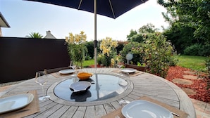 A round outdoor dining table with glass top, surrounded by plates, bowls, and cutlery, set on a wooden deck with a pergola overhead.