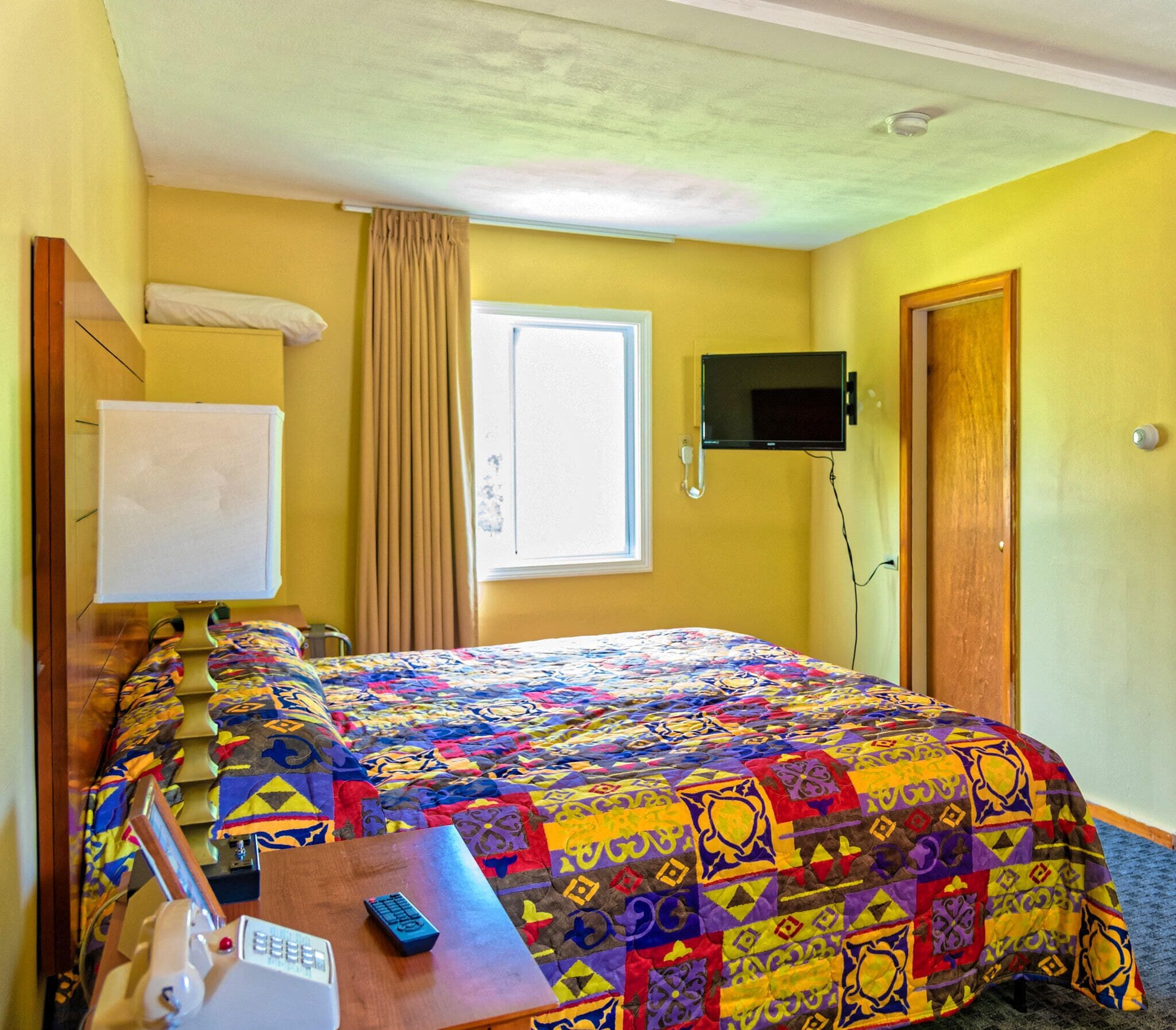 A hotel room with a bed, a desk with a computer and a remote control, a TV mounted on the wall, and a window with curtains.