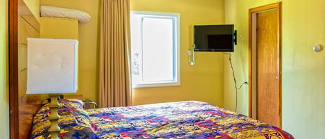 A hotel room with a bed, a desk with a computer and a remote control, a TV mounted on the wall, and a window with curtains.