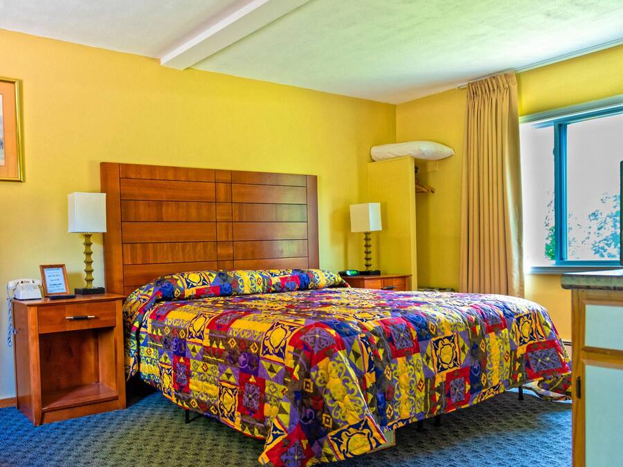 A hotel room with a wooden headboard, a colorful quilted bedspread, two bedside lamps, a desk with a telephone, and a window with curtains.