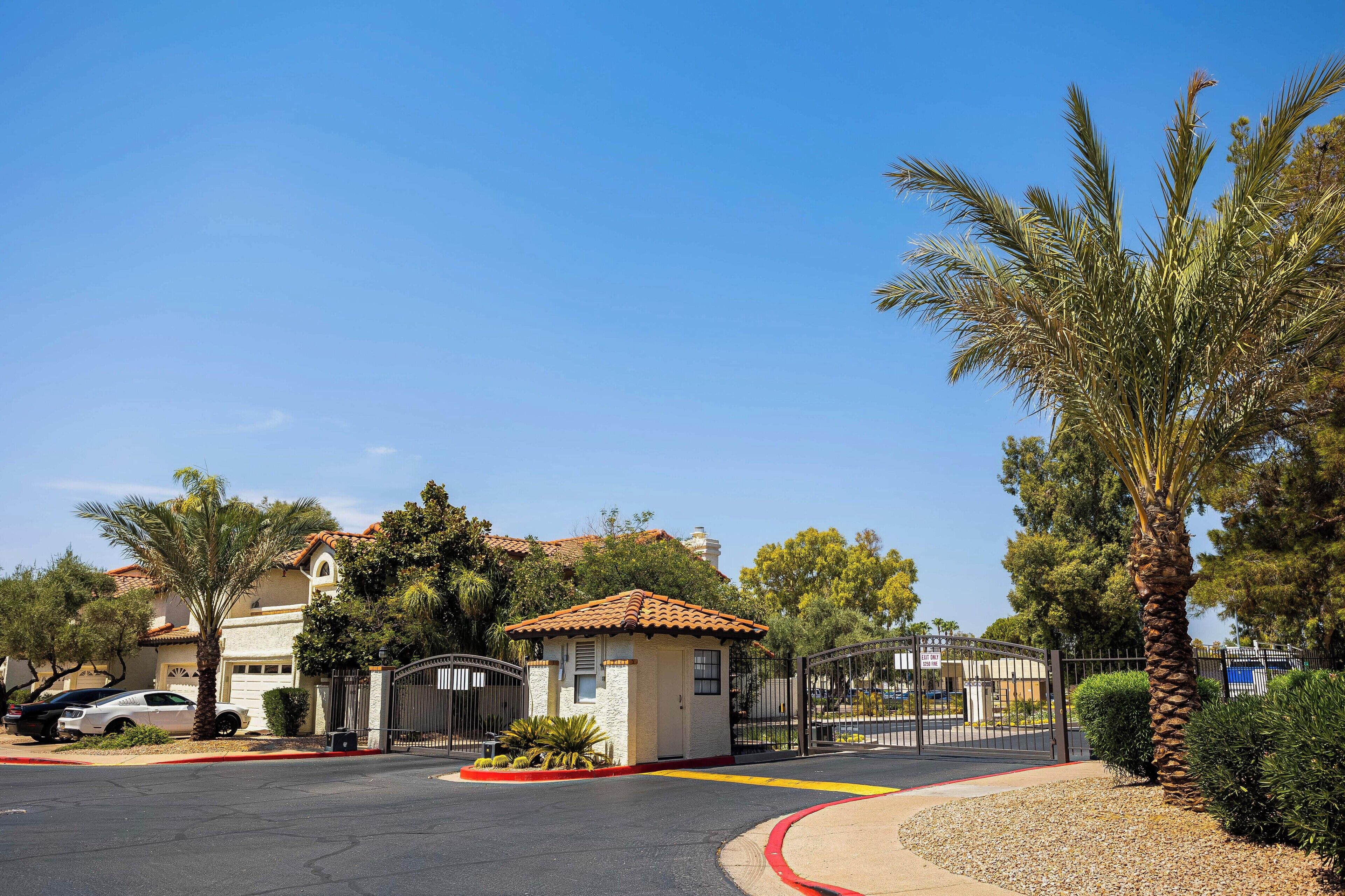 A gated residential area with a small building, palm trees, and a clear blue sky.
