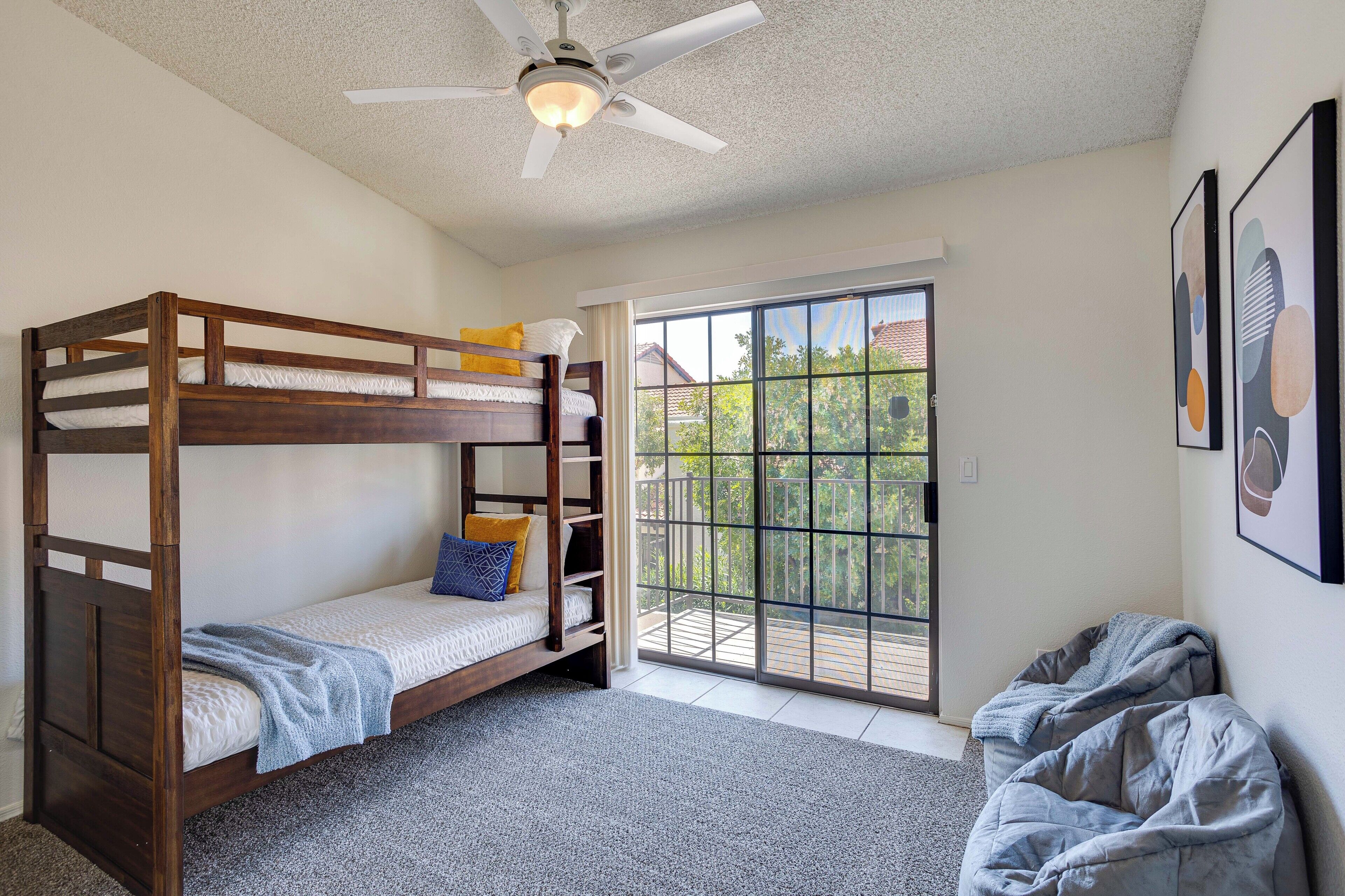A room with a bunk bed, a ceiling fan, a large window with a view of greenery, and a balcony.