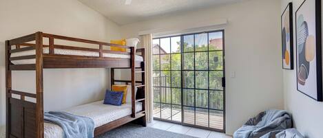 A room with a bunk bed, a ceiling fan, a large window with a view of greenery, and a balcony.