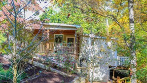 A rustic cabin with a wooden deck, surrounded by lush greenery and a stone wall.