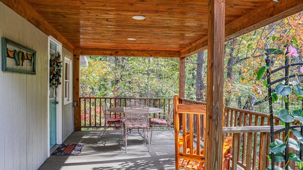 A covered porch with a wooden ceiling, a table with chairs, and a view of autumn foliage.