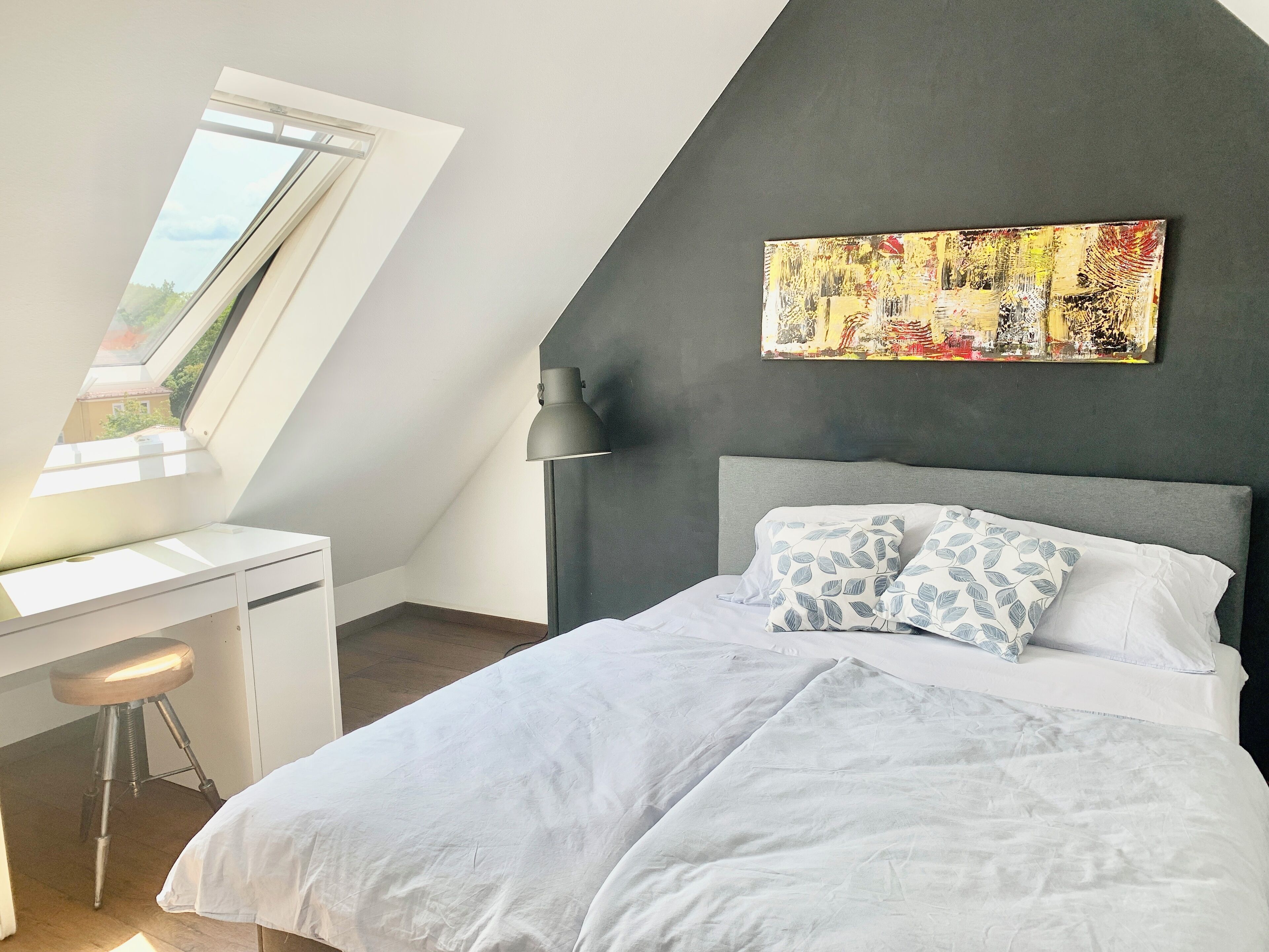A bedroom with a sloped ceiling, a desk, a bed with patterned pillows, and a large abstract painting on the wall.