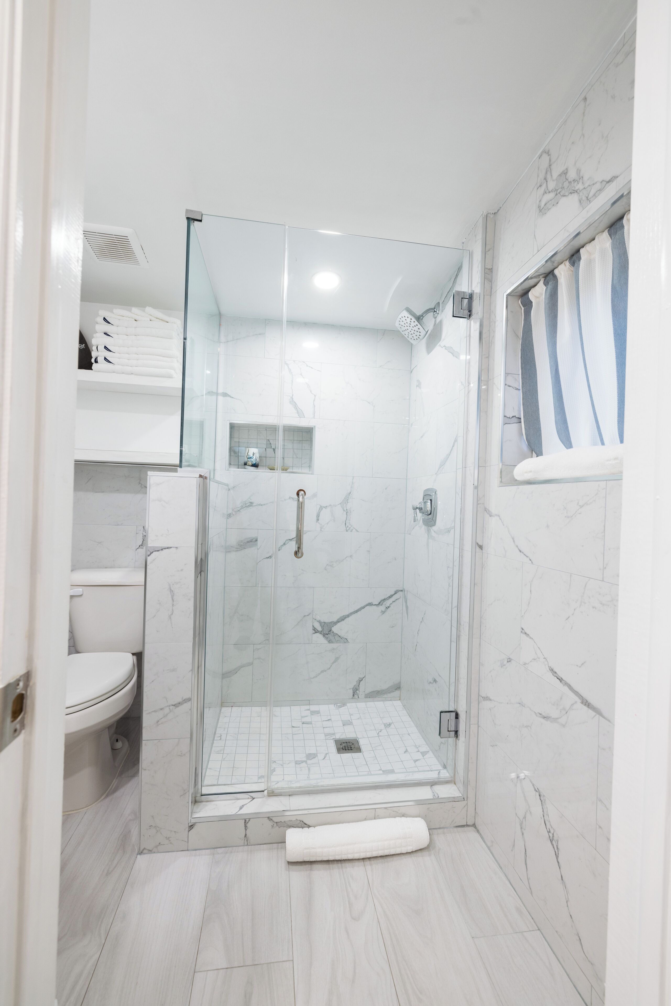 Un baño moderno con ducha de vidrio, azulejos de mármol blanco, inodoro y toallero.