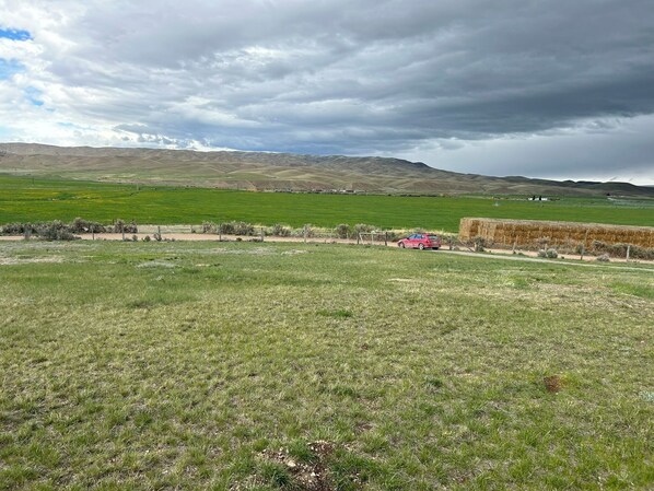 A rural landscape with a red car parked on a dirt path, surrounded by green fields and distant hills under a cloudy sky.