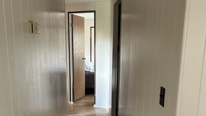 A narrow hallway with wooden flooring and white walls, leading to a bedroom with a bed and a door. - Cozy Quiet Home on Sage Creek (Dell)