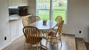 A dining area with a round table and four chairs, a kitchen with a built-in shelf, and a large window with a view of a grassy yard. - Cozy Quiet Home on Sage Creek (Dell)