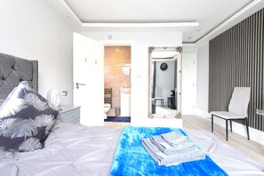 A modern bedroom with a large bed, a bedside table, a chair, and a view into the bathroom.