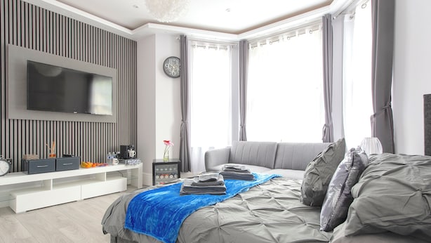 A modern bedroom with a large bed, a flat-screen TV mounted on the wall, a bedside table with a lamp, and a window with curtains. - The Pond House - Ensuite Rooms (London)
