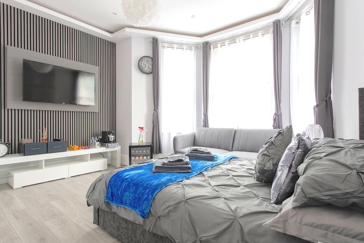 A modern bedroom with a large bed, a flat-screen TV mounted on the wall, a bedside table with a lamp, and a window with curtains.