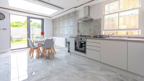 A modern kitchen with a large island, a dining area with chairs, and a view of the garden through a glass door. - The Pond House - Ensuite Rooms (London)