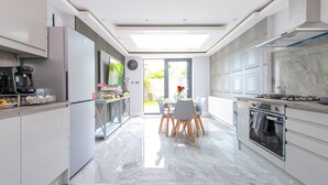 A modern kitchen with a central island, built-in appliances, and a dining area with a view to the outdoors. - The Pond House - Ensuite Rooms (London)
