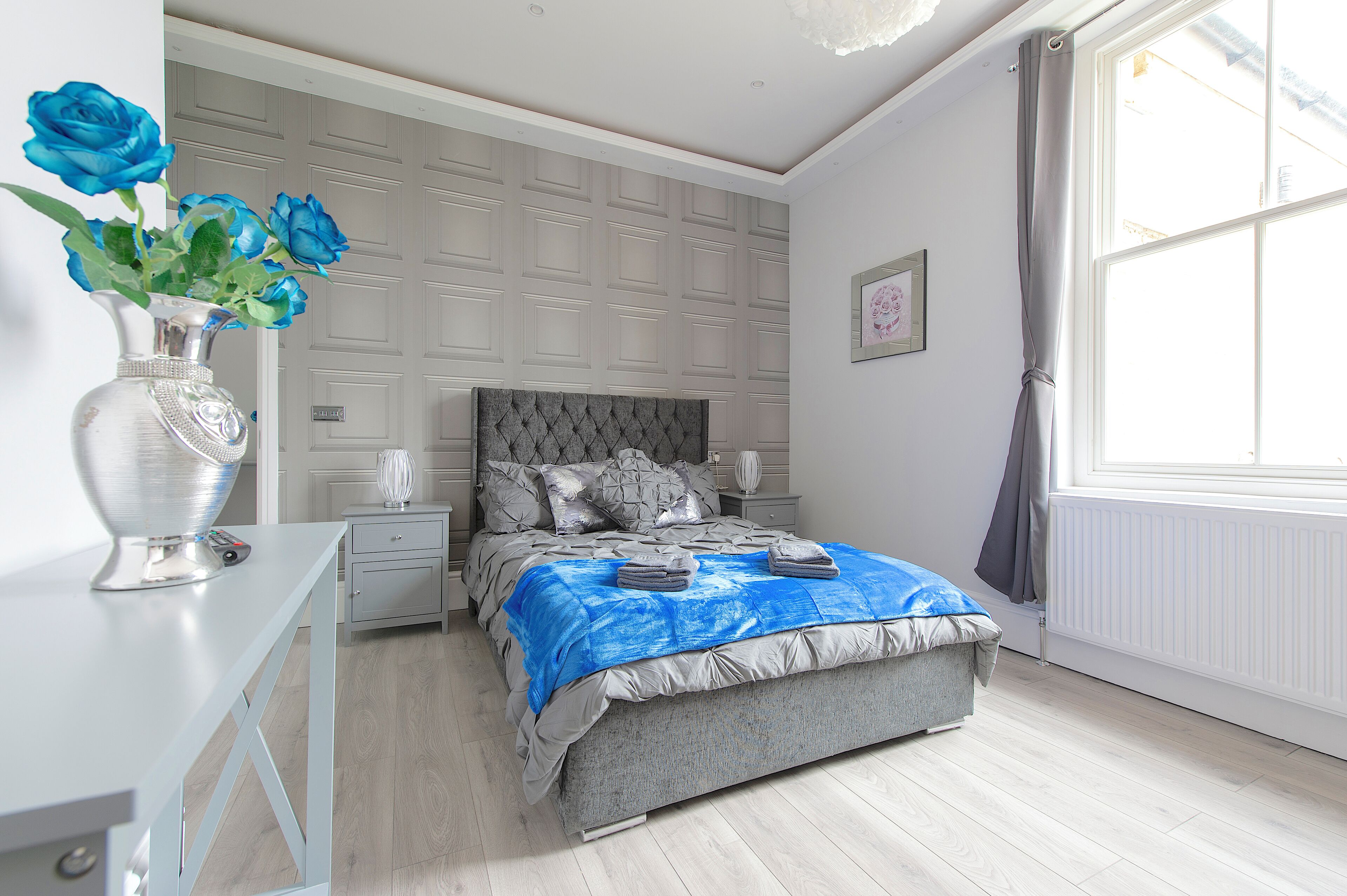 A bedroom with a grey bed, a white dresser, a vase with blue flowers, and a window with curtains.