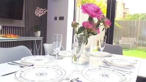 A dining table set with white plates, silverware, and champagne flutes, with a vase of pink flowers in the center. - The Pond House - Ensuite Rooms (London)