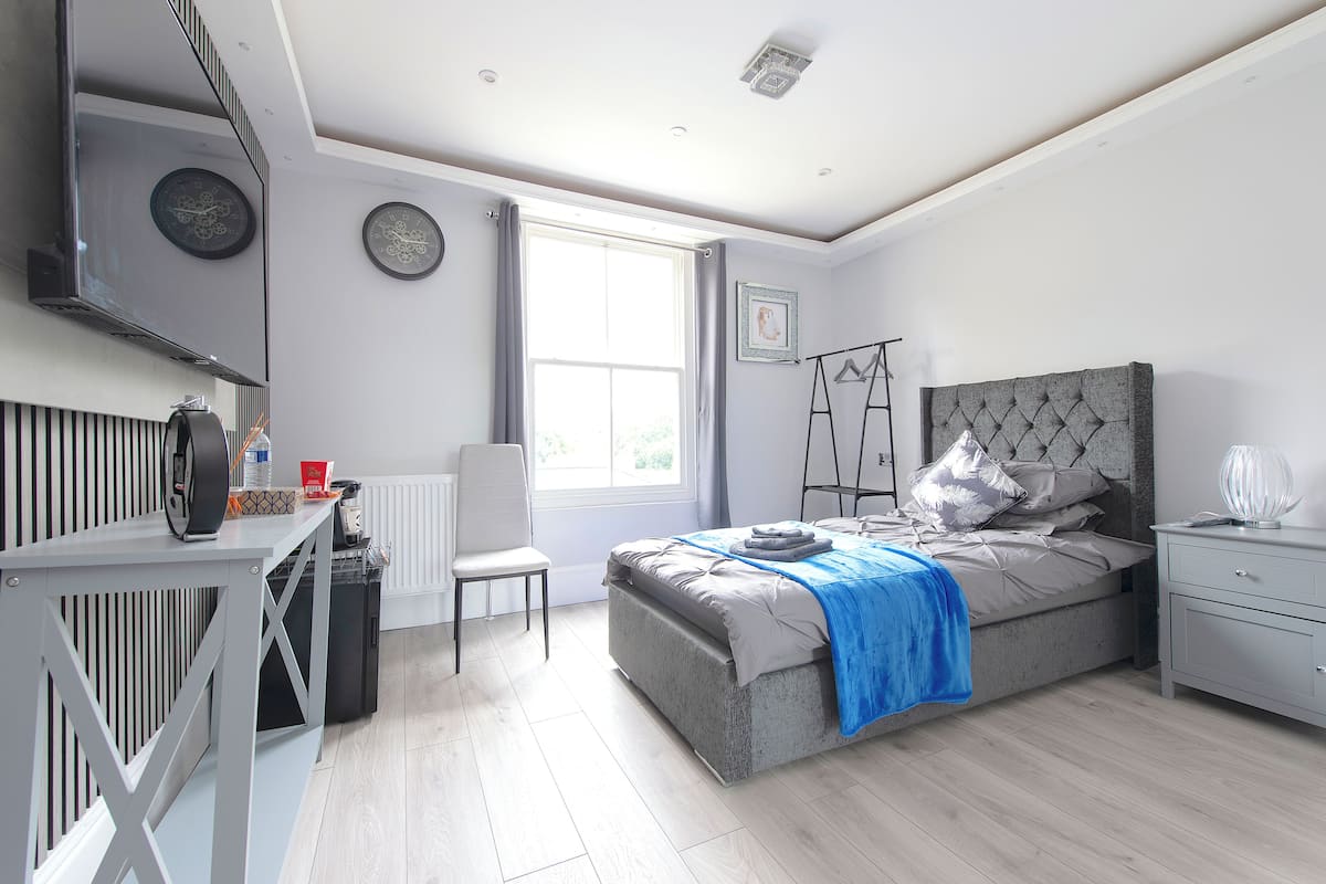 A bedroom with a bed, bedside table, chair, and a desk with a chair. There is a window with curtains, a radiator, and a wall-mounted clock.
