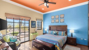 A bedroom with a large bed, a ceiling fan, a window with a view, and a nightstand with a lamp.
