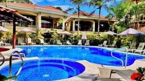 Una piscina amb gandules i ombrel·les, envoltada d'un edifici amb teulada enrajolada i vegetació.