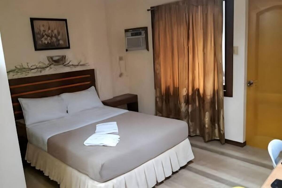 A hotel room with a bed, a nightstand, a window with curtains, and a door.