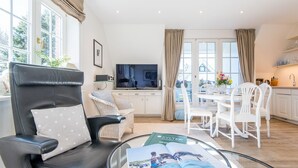 A modern living room with a leather chair, a glass coffee table, a dining area with white chairs, a kitchen with white cabinets, and a large window with curtains. - Haus Skandia, App. 4 - Strandnahe 3-zimmer Ferienwohnung auf ca. 56 m² Wohnfläche, für bis zu 4 (Wenningstedt-Braderup (Sylt))