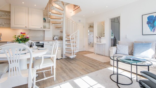 A modern kitchen with white cabinets, a dining area with white chairs, a spiral staircase, and a living room with a sofa and a coffee table. - Haus Skandia, App. 4 - Strandnahe 3-zimmer Ferienwohnung auf ca. 56 m² Wohnfläche, für bis zu 4 (Wenningstedt-Braderup (Sylt))