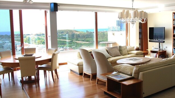 A modern living room with a large window, a sofa, a dining table, and a chandelier.