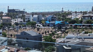 A coastal residential area with modern buildings, a clear view of the sea, and a well-maintained public space with trees and umbrellas.