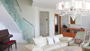 A modern living room with a glass staircase, a piano, a beige sofa, and a chandelier.