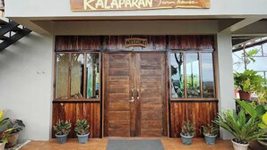 Una puerta de madera con un cartel que dice 'Welcome' encima, flanqueada por macetas con plantas, que da paso a un edificio con un cartel que indica 'Kalaparang Farmhouse'.